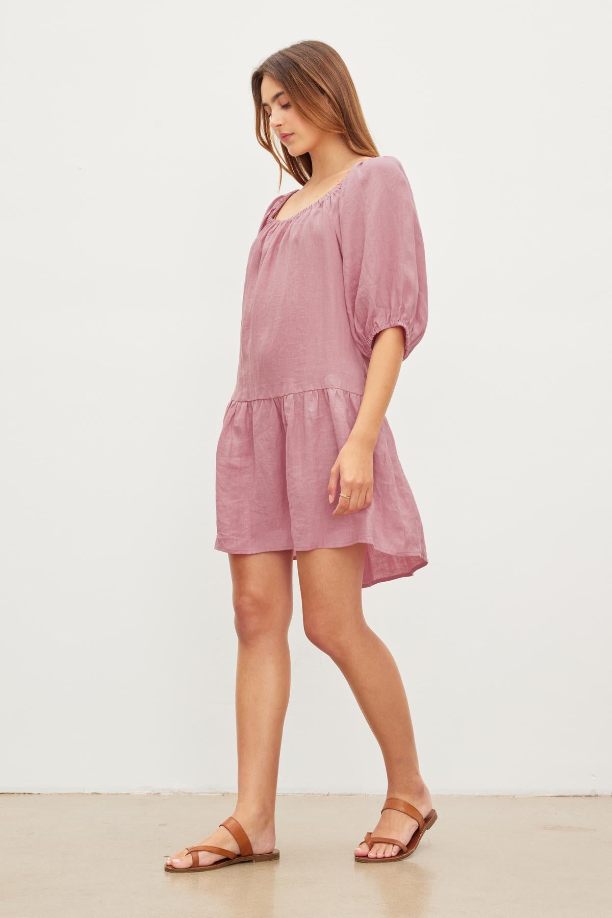 A woman in a pink Velvet by Graham & Spencer IRINA LINEN TIERED DRESS and brown sandals standing with her head tilted down, against a plain white background.-36571205402817