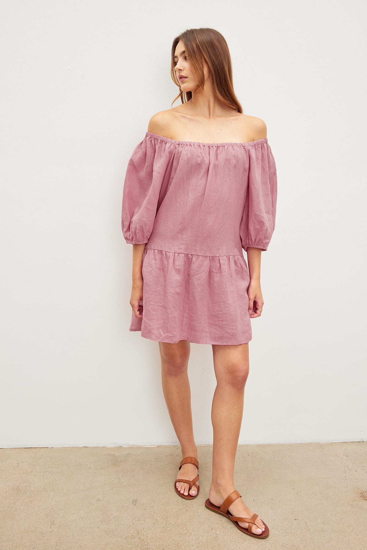   A person stands against a plain background wearing the IRINA LINEN DRESS by Velvet by Graham & Spencer, an off-shoulder pink dress with three-quarter sleeves, an elastic neckline, and inseam pockets, paired with brown sandals. 