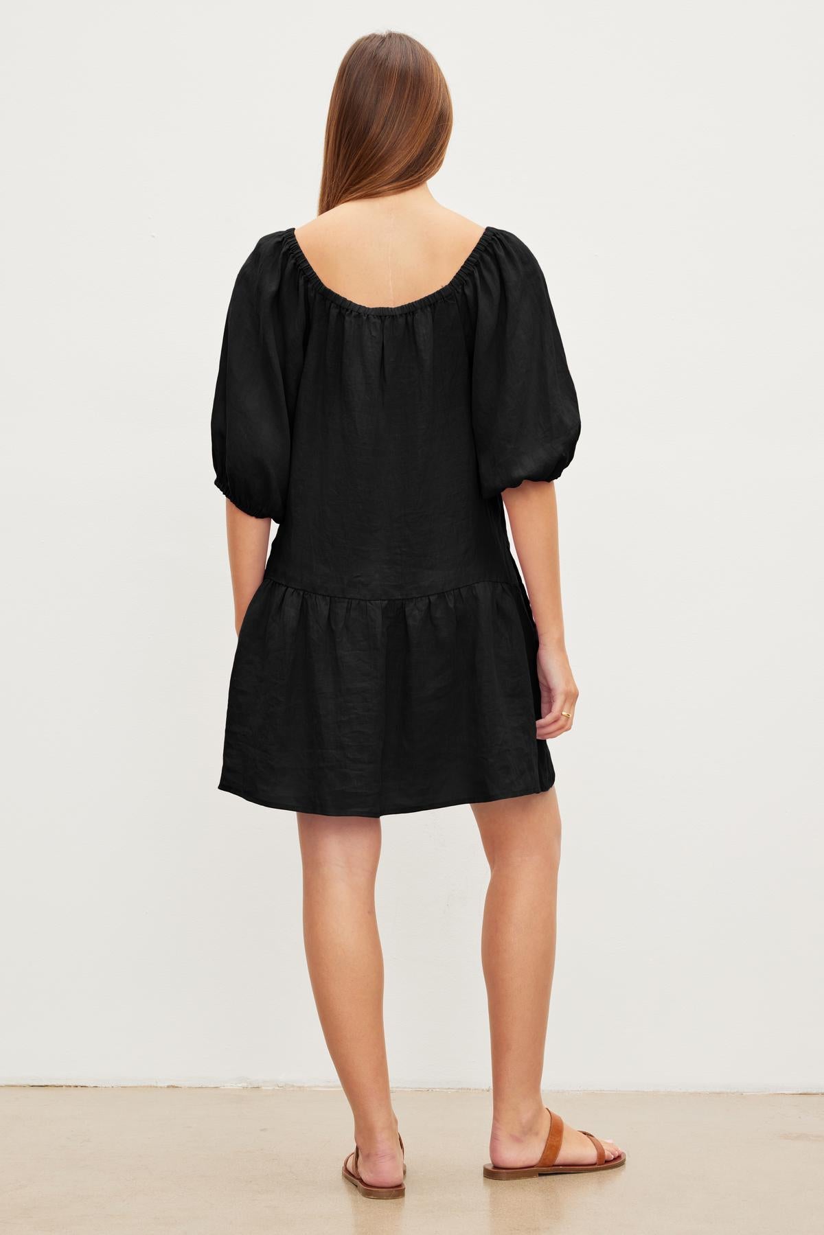 Woman standing with her back to the camera, wearing a black short-sleeved IRINA LINEN DRESS by Velvet by Graham & Spencer and brown sandals.-37229080969409