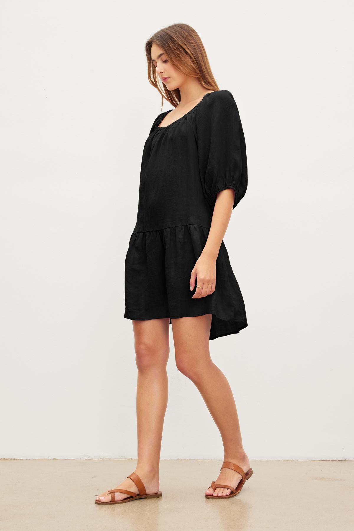   A woman in a loose black IRINA LINEN DRESS by Velvet by Graham & Spencer with inseam pockets and brown sandals stands against a plain white background, looking downwards. 