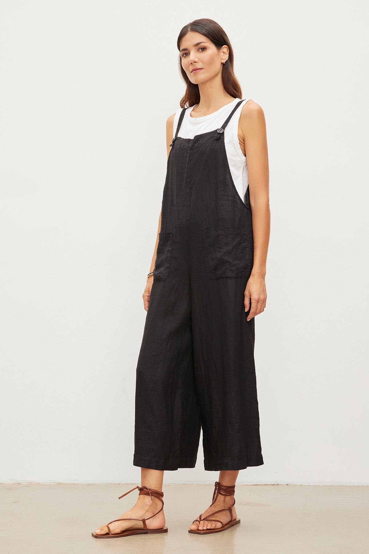 Woman standing in a studio wearing black overalls with patch pockets and a white tank top, paired with brown strappy sandals. - Woman standing in a studio wearing Velvet by Graham & Spencer's Isabel Linen Jumper, paired with brown strappy sandals.-35967680151745