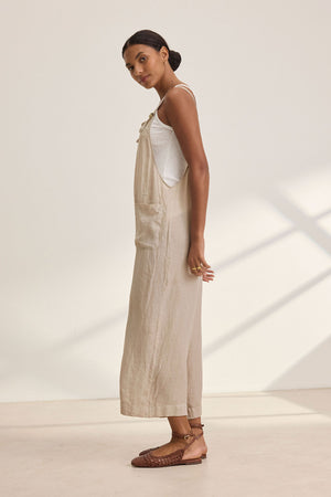 A woman stands sideways in a white tank top with the ISABEL LINEN ROMPER by Velvet by Graham & Spencer and brown woven sandals, showcasing a relaxed and versatile style perfect for warm weather, all set against a crisp white backdrop.