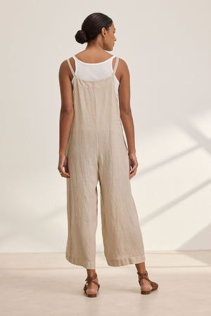 A woman faces away from the camera, modeling the ISABEL LINEN ROMPER by Velvet by Graham & Spencer in beige over a white top with brown sandals, reflecting a relaxed and versatile style.
