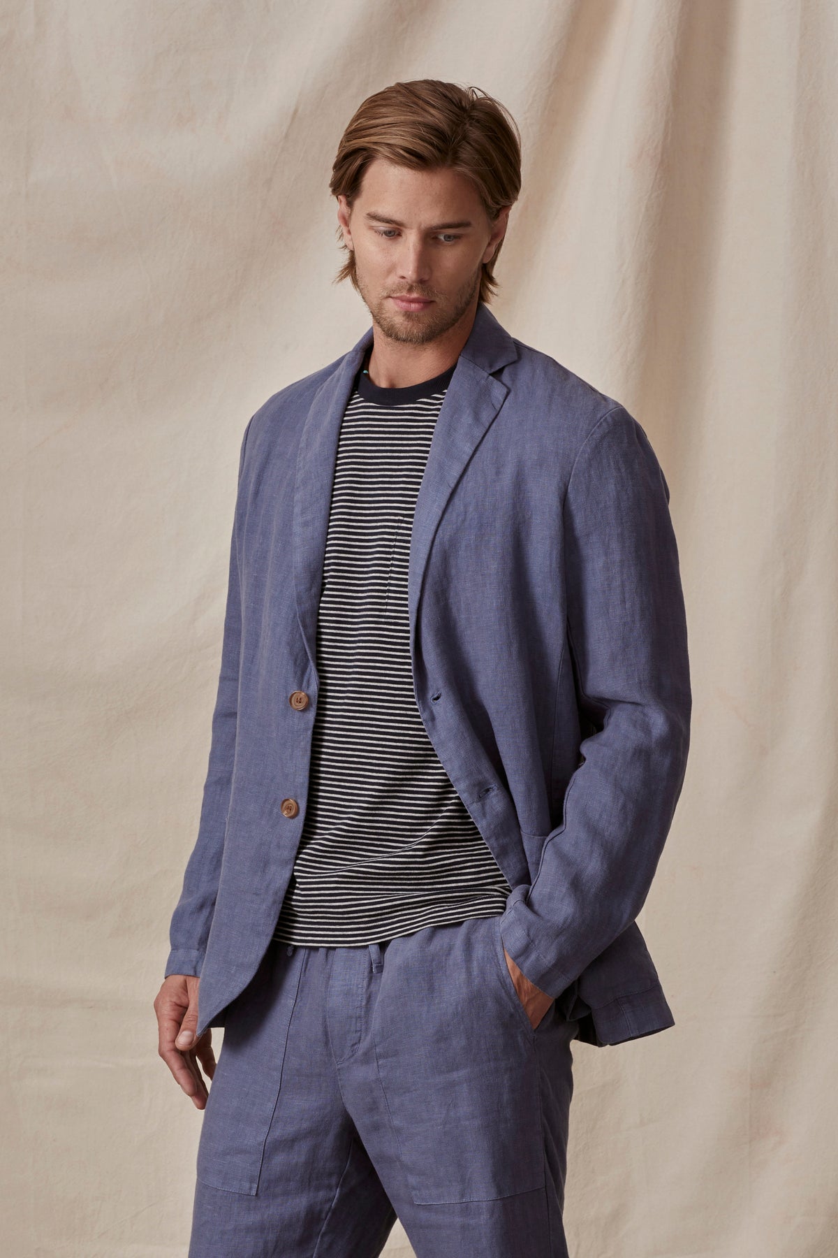 A man in a stylish blue Velvet by Graham & Spencer Joshua linen blazer and striped shirt, standing with hands in pockets against a neutral backdrop.-36805484839105