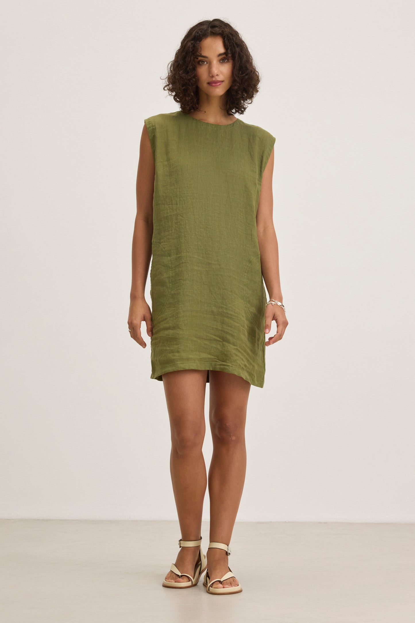 A woman stands in a plain room wearing the LARA LINEN DRESS by Velvet by Graham & Spencer, featuring a sleeveless design and detachable belt, paired with light-colored sandals and looking straight ahead with a neutral expression.-39274374103233