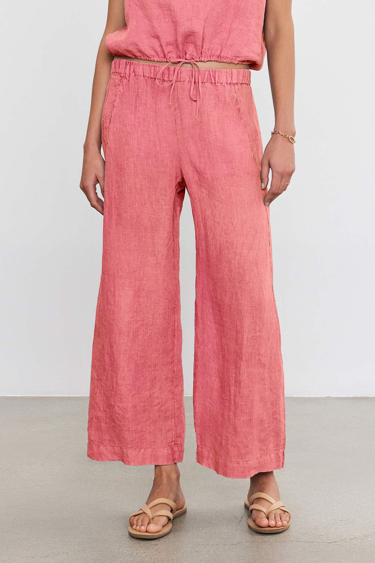   A person wearing lightweight pink LOLA LINEN PANT by Velvet by Graham & Spencer with an elastic waistband and drawstring, a matching top, a bracelet on the left wrist, and beige sandals on a plain grey floor. 