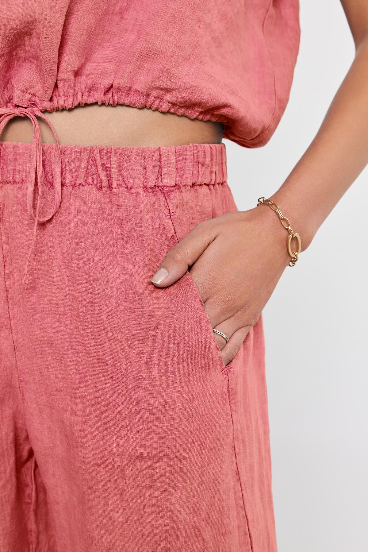   A person wearing a pink outfit with Velvet by Graham & Spencer's LOLA LINEN PANTs that feature a drawstring waist holds their right hand in their pocket, showing a gold bracelet and a ring on the same hand. 