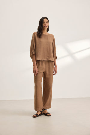 A person stands against a plain background wearing Velvet by Graham & Spencer's LOLA LINEN PANT—a relaxed-leg trouser with an elastic waist—paired with a brown long-sleeve top and black sandals.