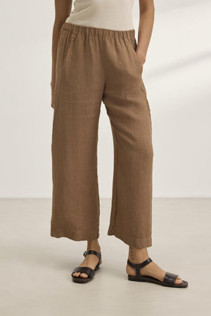 A person wearing Velvet by Graham & Spencer's LOLA LINEN PANT in brown and black sandals stands indoors on a light floor.