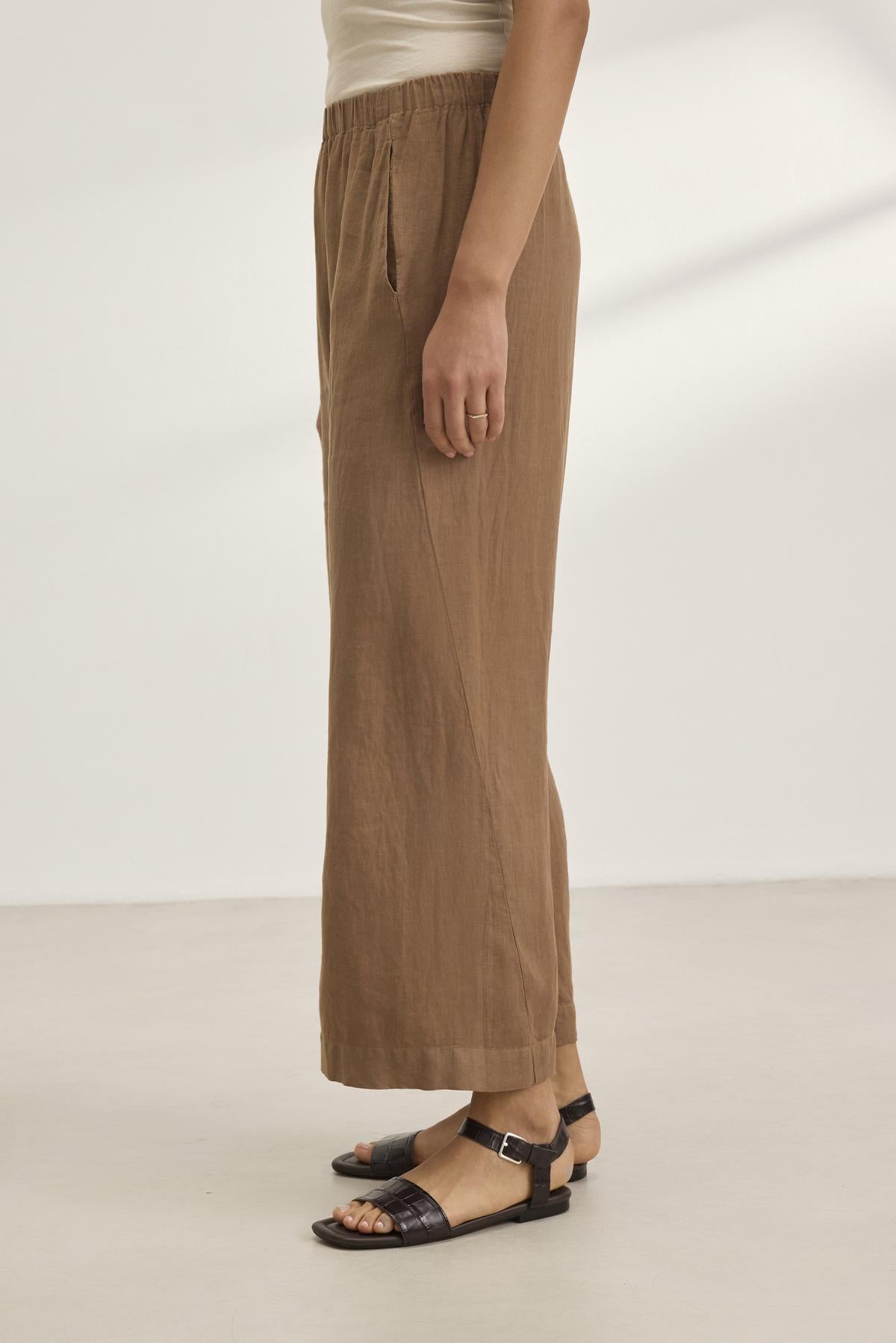 A person stands against a plain background, wearing Velvet by Graham & Spencer's LOLA LINEN PANT, a lightweight brown pull-on wide-leg pant with an elastic waist for comfort and style, paired with black sandals.-38657927315649