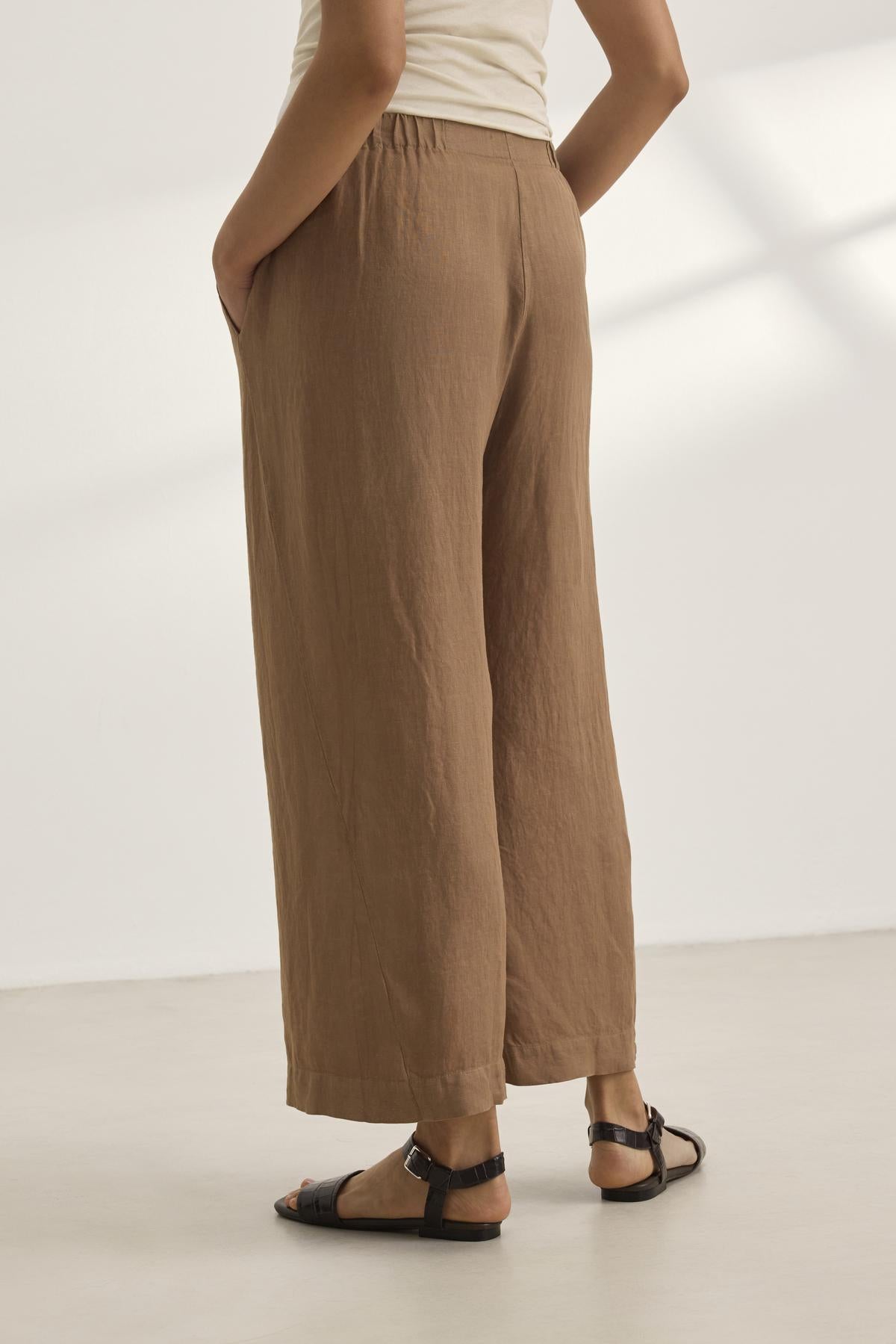 A person in a beige top and LOLA LINEN PANT from Velvet by Graham & Spencer—brown, wide-leg trousers with an elastic waist—paired with black sandals, stands on a light surface with hands in pockets.-38657927348417