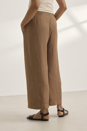 A person in a beige top and LOLA LINEN PANT from Velvet by Graham & Spencer—brown, wide-leg trousers with an elastic waist—paired with black sandals, stands on a light surface with hands in pockets.