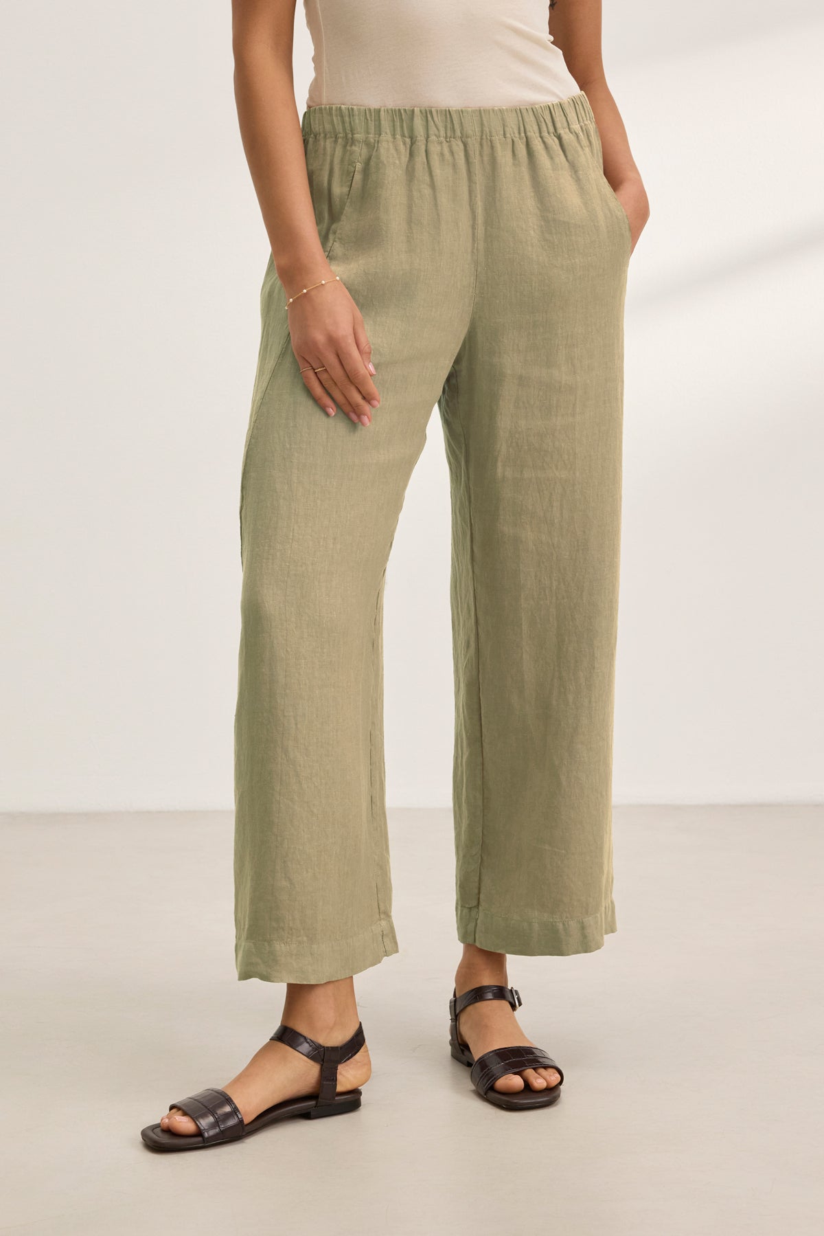 Wearing the Velvet by Graham & Spencer LOLA LINEN PANT in light olive green, a beige top, and black sandals, a person stands on a light floor with one hand in their pocket.-39359713116353