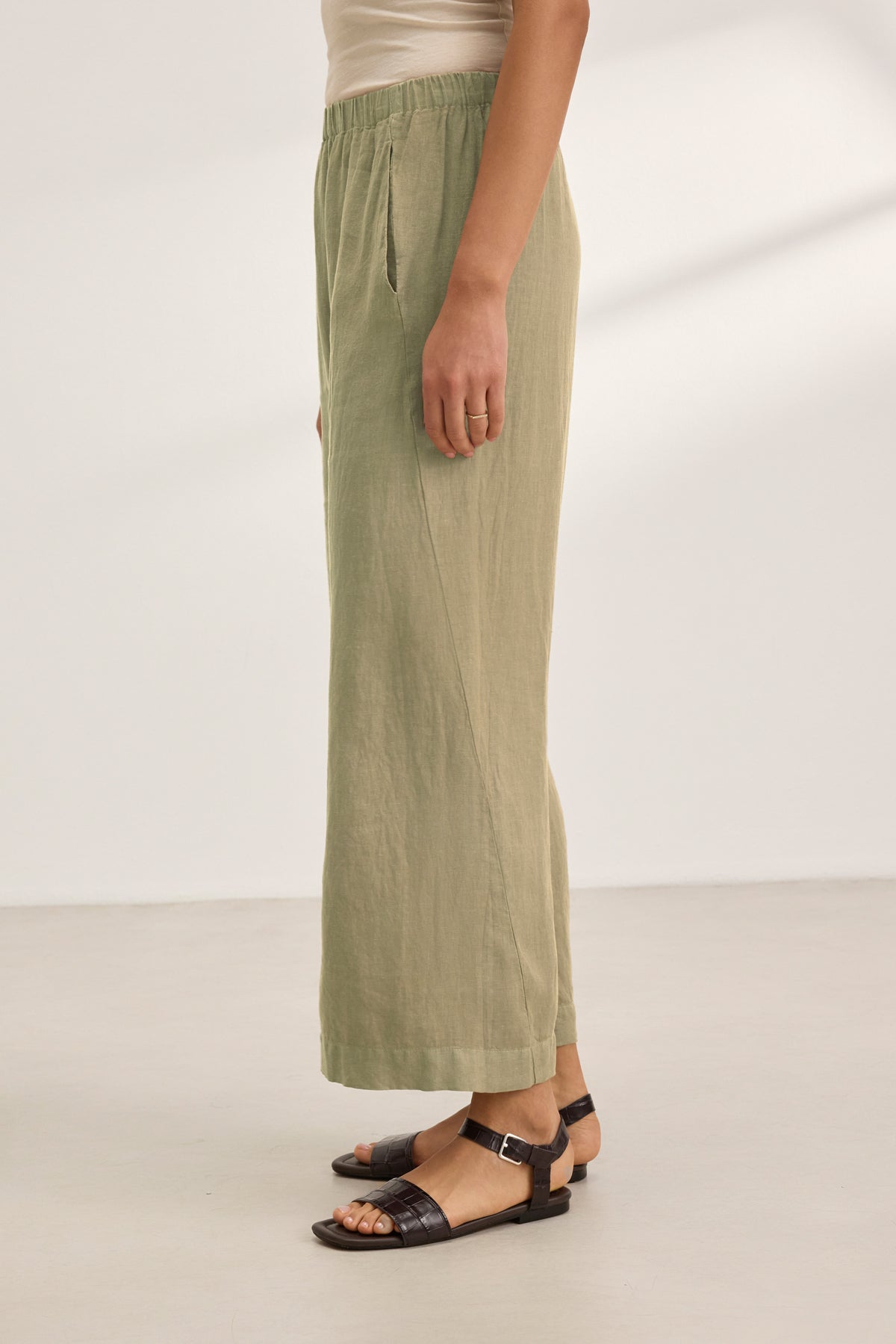 A person stands indoors wearing Velvet by Graham & Spencer's LOLA LINEN PANT—olive green, wide-leg pull-on trousers with an elastic waist. Only the lower body is visible; they are also wearing a beige top and dark brown sandals.-39359713149121