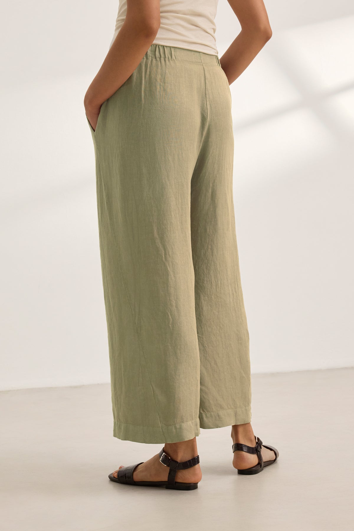 Person wearing the LOLA LINEN PANT by Velvet by Graham & Spencer in light olive green with an elastic waist, a sleeveless beige top, and black sandals, standing indoors with hands in pockets.-39359713181889