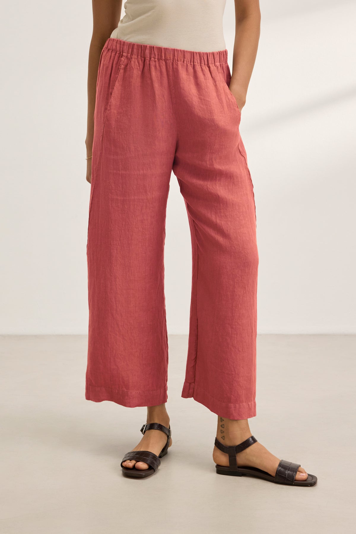 A person wears the LOLA LINEN PANT by Velvet by Graham & Spencer—loose, high-waisted, rose-colored linen pull-on trousers with an elastic waist—paired with a beige top and black sandals, standing on a light floor against a white background.-39395663970497
