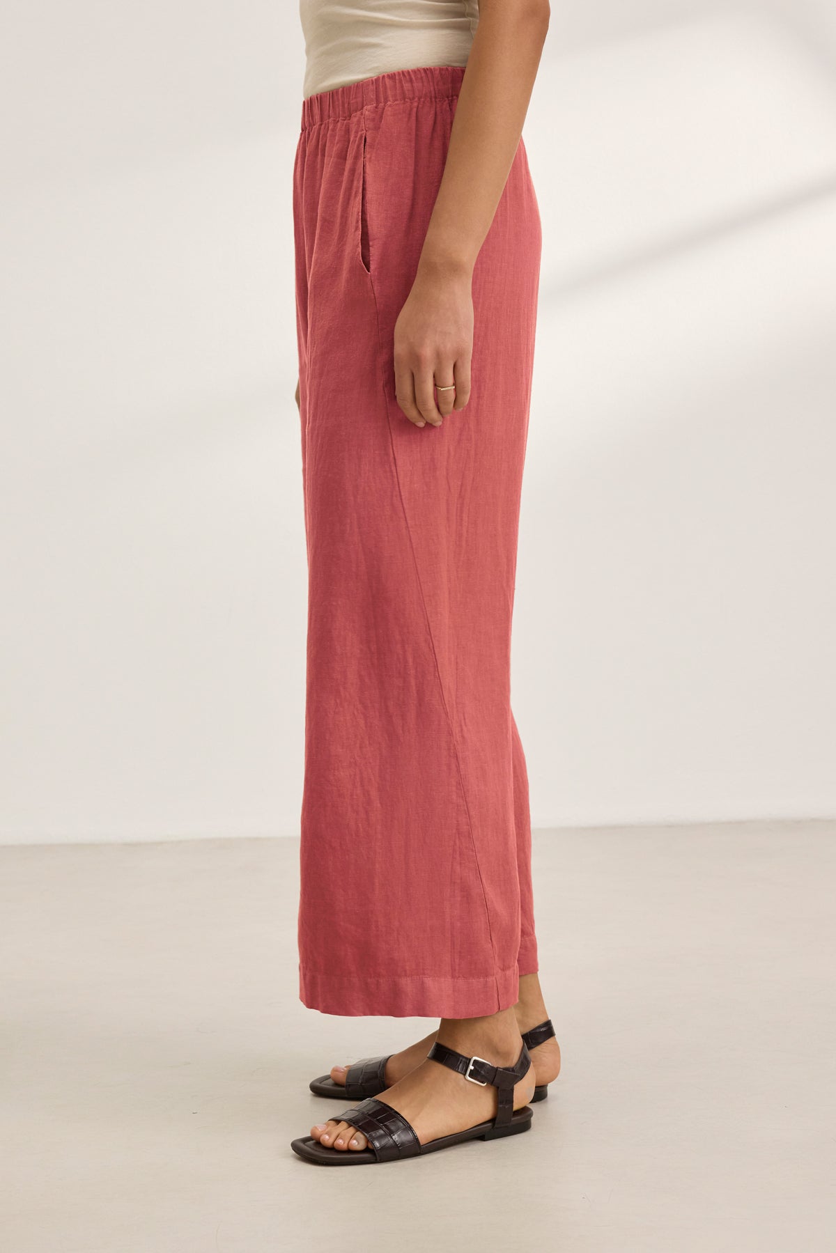 A person wearing the LOLA LINEN PANT by Velvet by Graham & Spencer in coral pink, paired with a beige top and black sandals, stands on a light-colored floor against a plain white background.-39395663904961