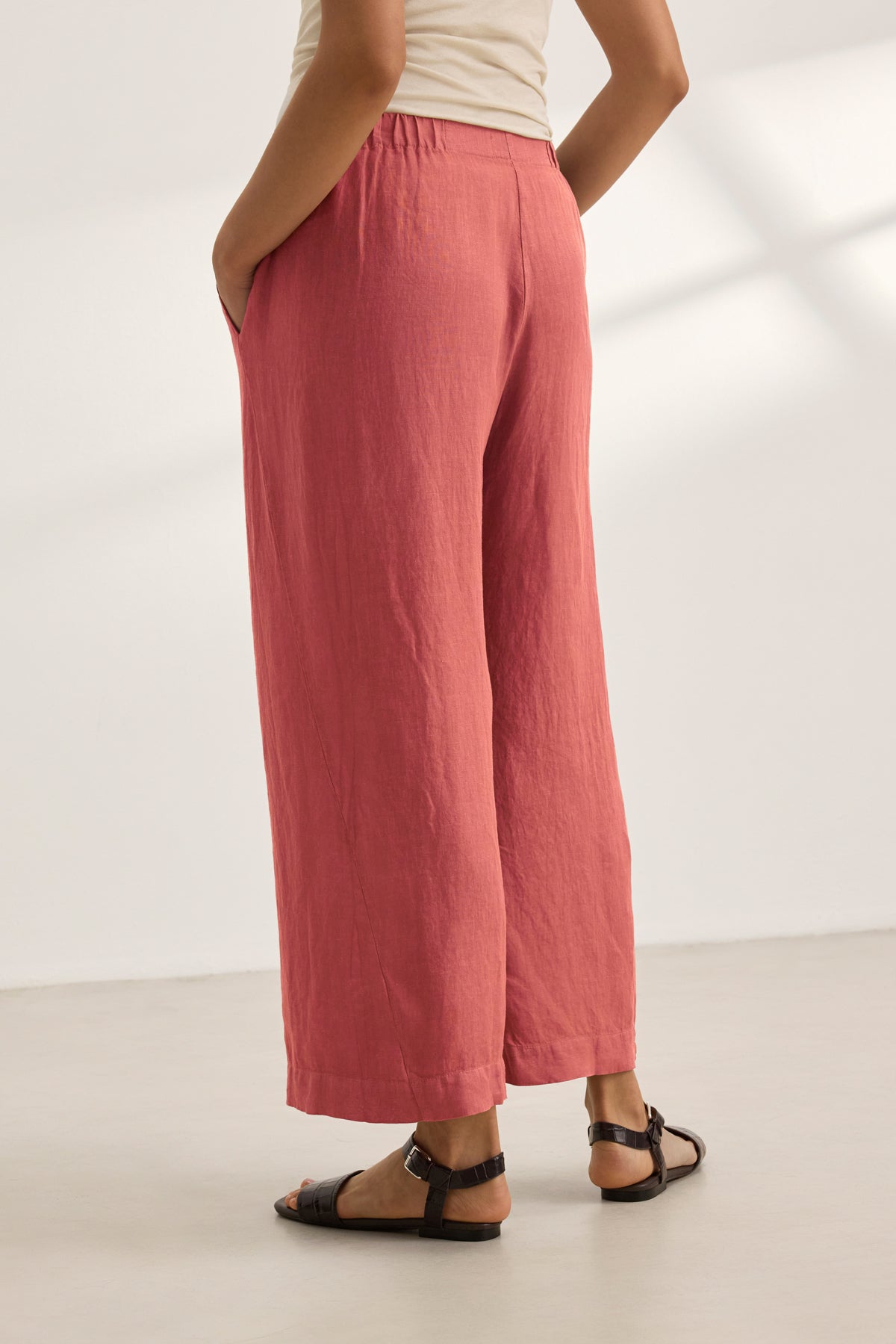 A person wears the LOLA LINEN PANT by Velvet by Graham & Spencer—loose, pink, pull-on trousers with an elastic waist—paired with a beige top and black sandals, standing against a plain white background, hands in pockets.-39395663872193