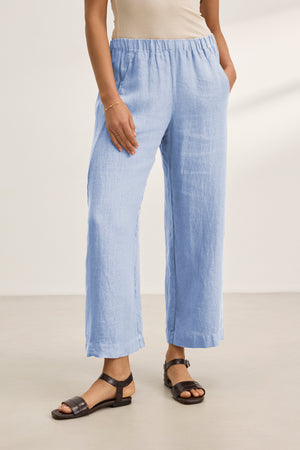 A person wears the LOLA LINEN PANT by Velvet by Graham & Spencer in light blue with a beige sleeveless top and black sandals, standing on a light floor, hands in pockets.