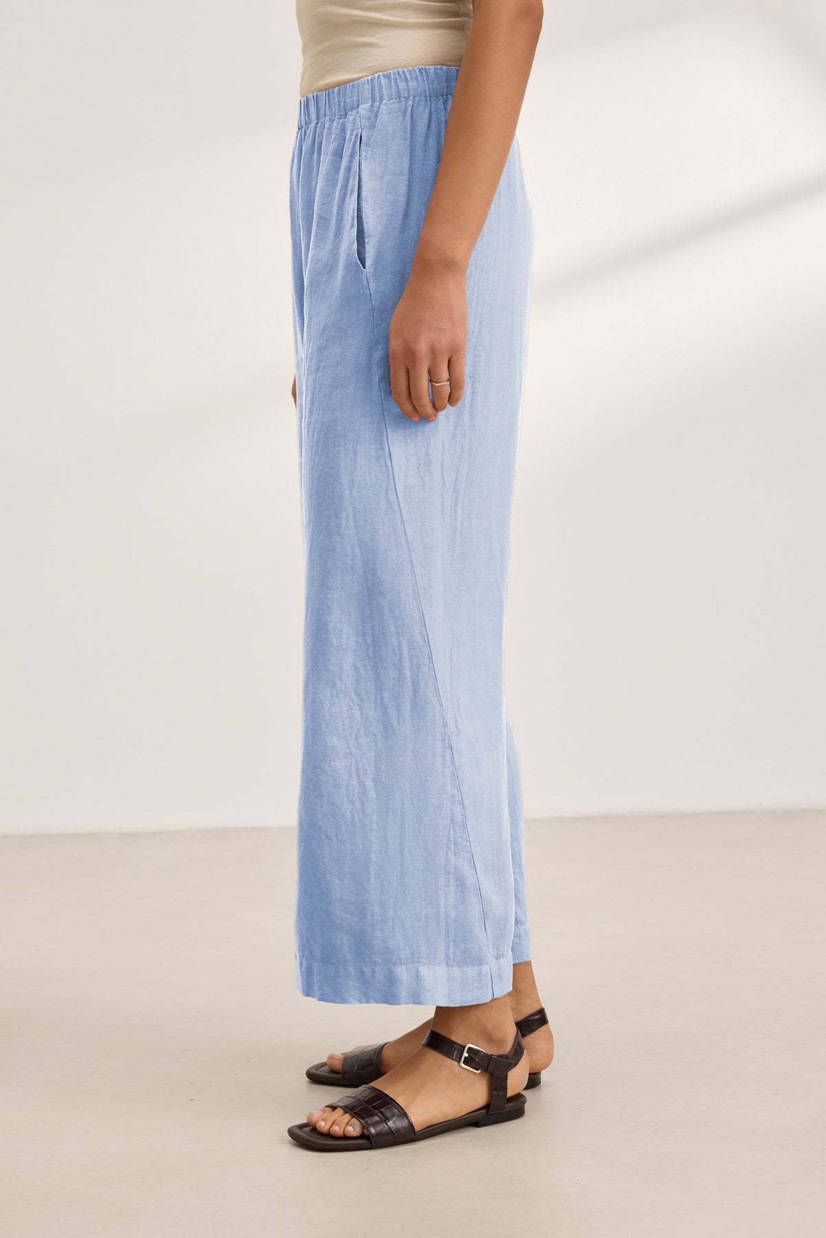   A person stands sideways against a plain light background, wearing Velvet by Graham & Spencer's LOLA LINEN PANT in light blue with an elastic waist, paired with a beige top and black sandals. 
