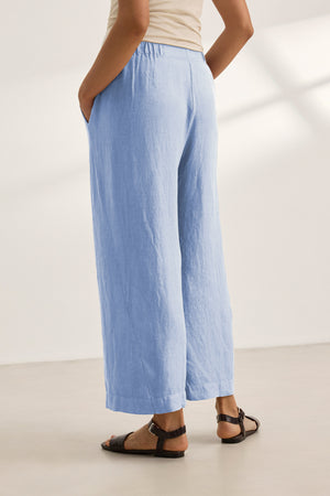 A person wears the LOLA LINEN PANT by Velvet by Graham & Spencer in light blue with an elastic waist, paired with a beige top and black sandals, standing indoors with hands in pockets.