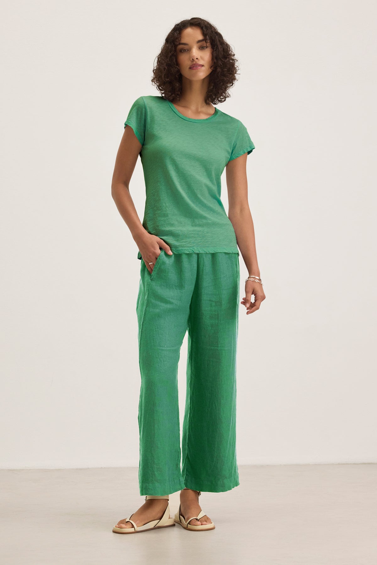 A woman stands indoors wearing the Velvet by Graham & Spencer ODELIA TEE in green, paired with matching wide-leg pants and beige sandals. She has one hand in her pocket and looks at the camera.-39395676061889