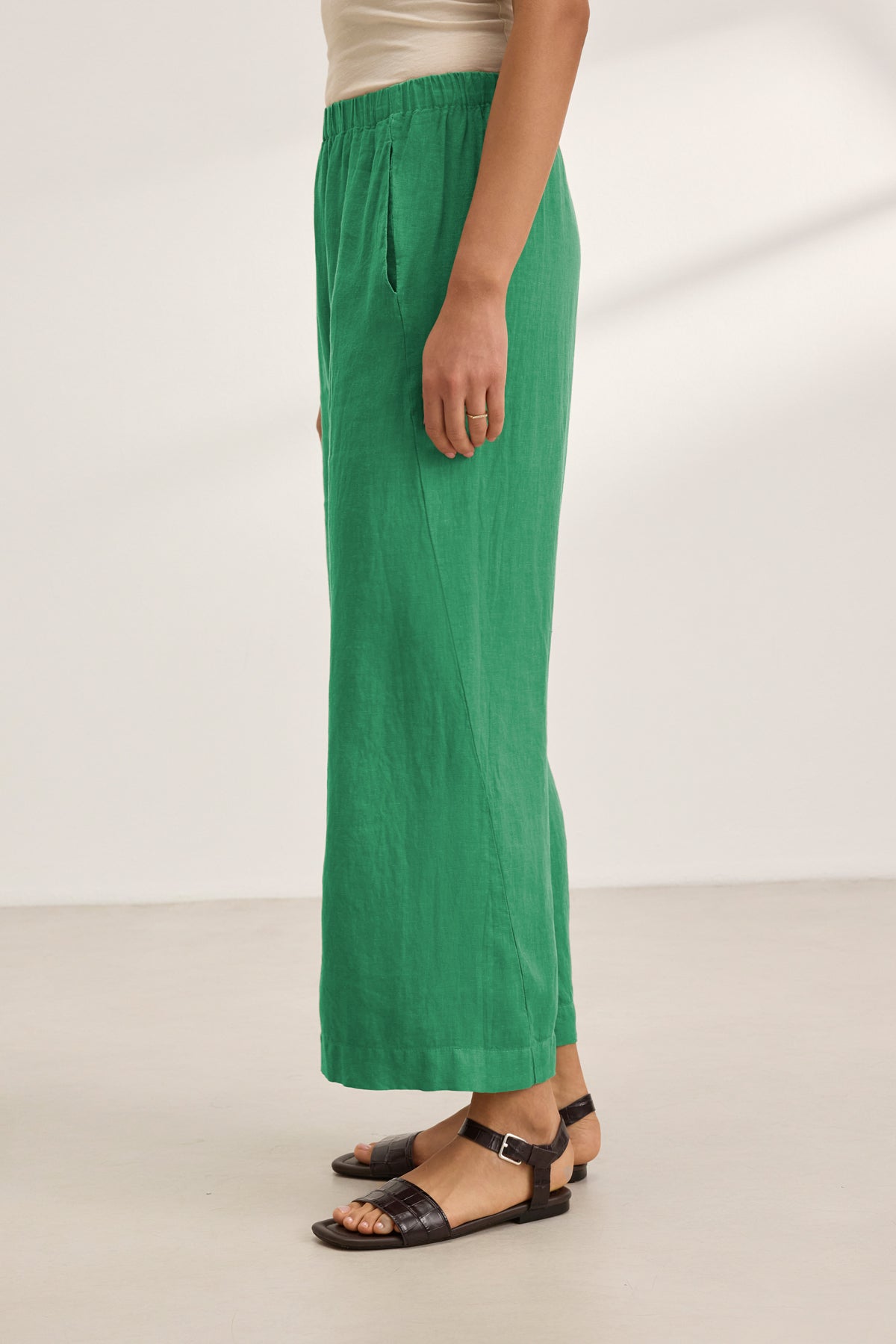 A person stands sideways on a light floor against a plain background, wearing the Velvet by Graham & Spencer LOLA LINEN PANT in green with an elastic waist and wide-leg fit, paired with a beige top and brown sandals.-39333752504513