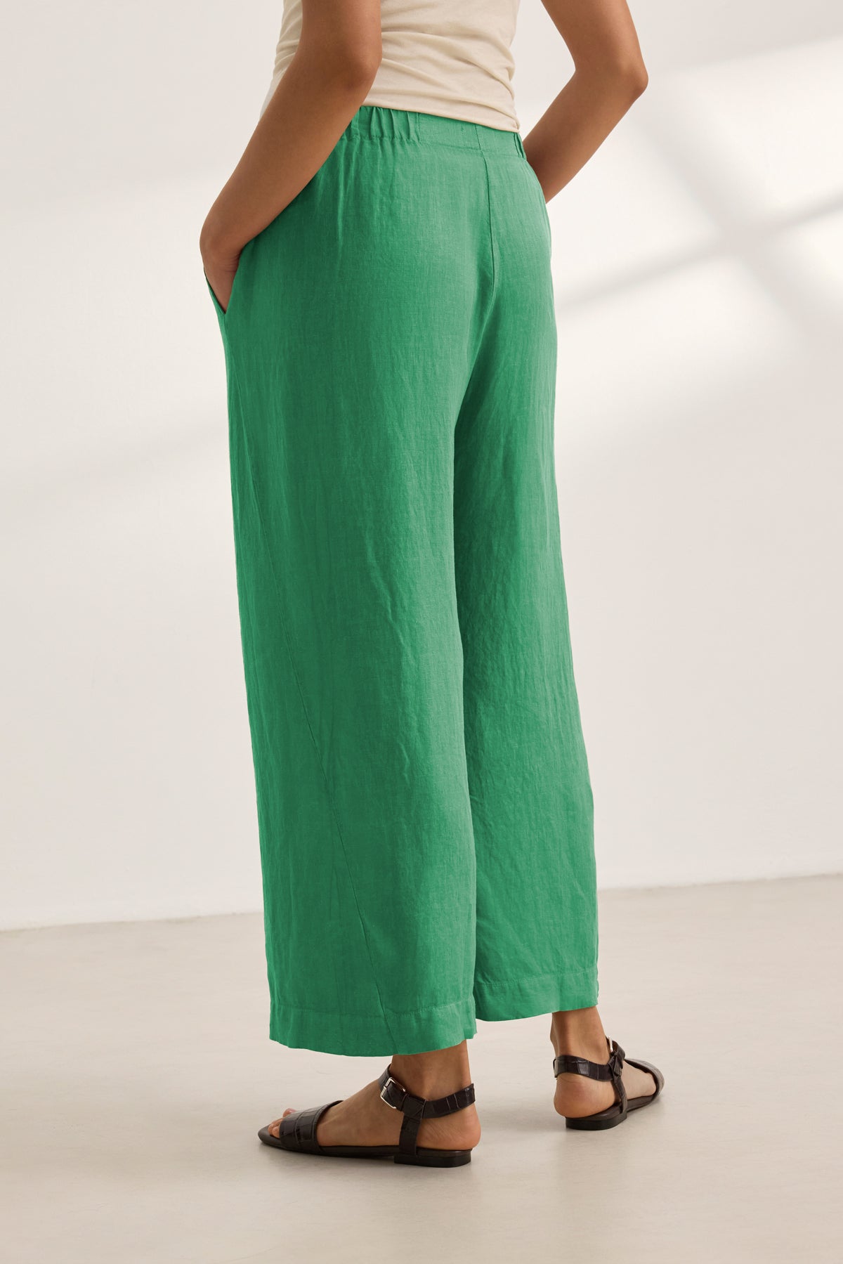   A person wears the LOLA LINEN PANT by Velvet by Graham & Spencer in green, paired with a beige top and black sandals, standing indoors with hands in pockets and facing away from the camera. 