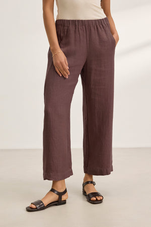 A person wearing a sleeveless beige top and Velvet by Graham & Spencer's LOLA LINEN PANT—lightweight brown cropped pants with an elastic waist and relaxed leg—plus black sandals, stands with one hand in their pocket.