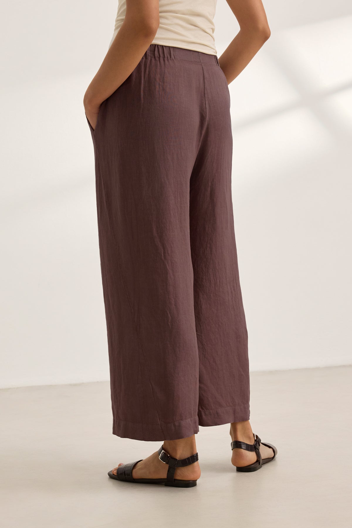   Model wearing the LOLA LINEN PANT by Velvet by Graham & Spencer in brown with black sandals, standing hands-in-pockets against a plain, light background. 