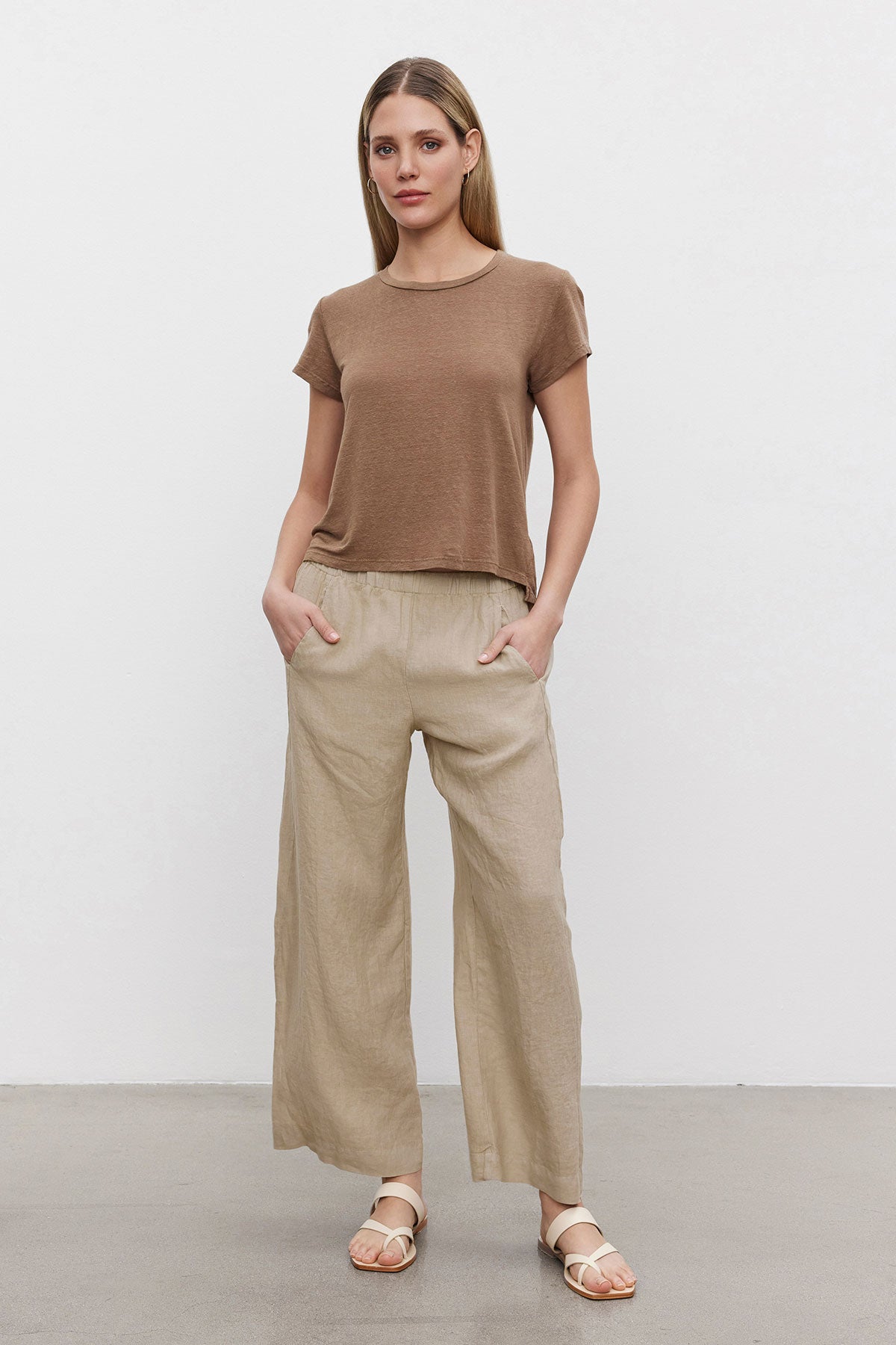   A woman stands in a neutral pose wearing a brown t-shirt, Velvet by Graham & Spencer LOLA LINEN PANTS with an elastic waist, and white sandals against a plain backdrop. 