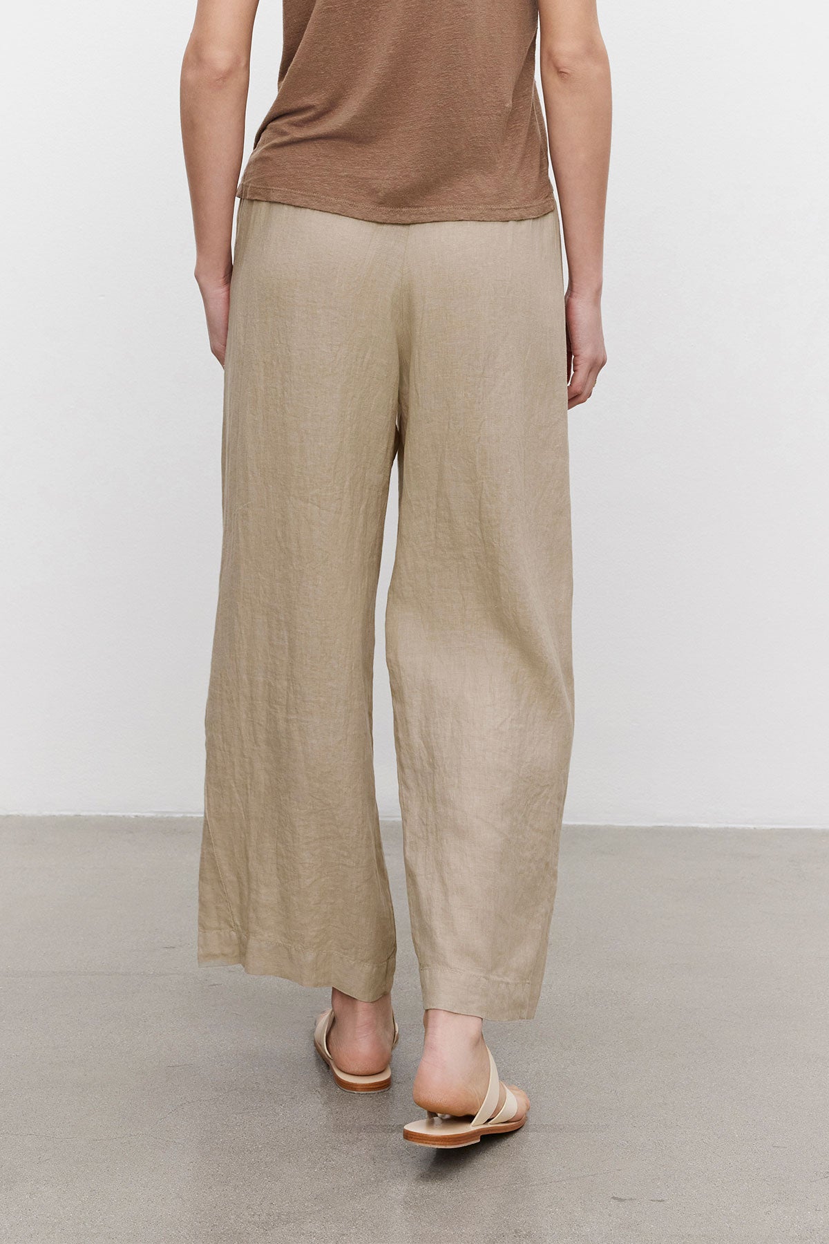   Rear view of a person wearing Velvet by Graham & Spencer LOLA LINEN PANTS and tan sandals, standing in a room with a white background. 