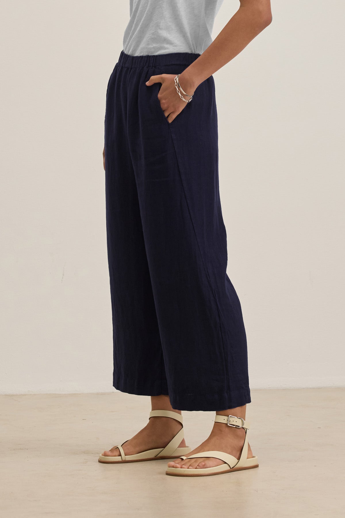 Person wearing a light gray t-shirt, white sandals, and Velvet by Graham & Spencer's LOLA LINEN PANT—wide-leg navy blue linen trousers with an elastic waist—stands on a light floor with one hand in their pocket.-41073344643265