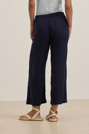 A person stands on a light floor facing away, wearing Velvet by Graham & Spencer's LOLA LINEN PANT in navy blue with an elastic waist, paired with a light gray top and beige sandals.