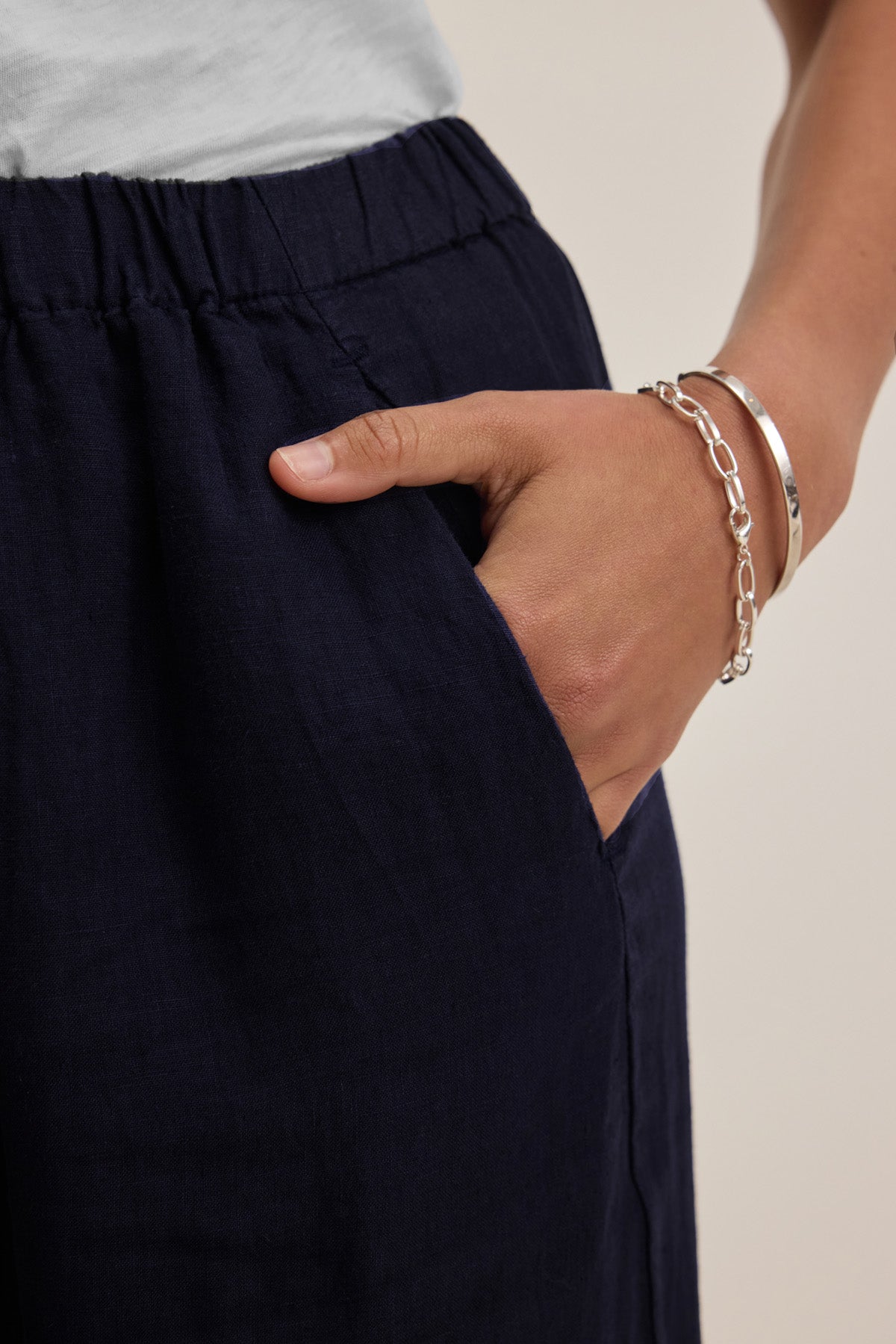 Someone wearing a white top and the LOLA LINEN PANT by Velvet by Graham & Spencer, styled with their right hand in their pocket, displaying a bracelet and chain on their wrist.-41073344577729