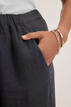 A person wearing the LOLA LINEN PANT by Velvet by Graham & Spencer with an elastic waist, paired with a white top and silver bracelets, has their right hand in their pocket.