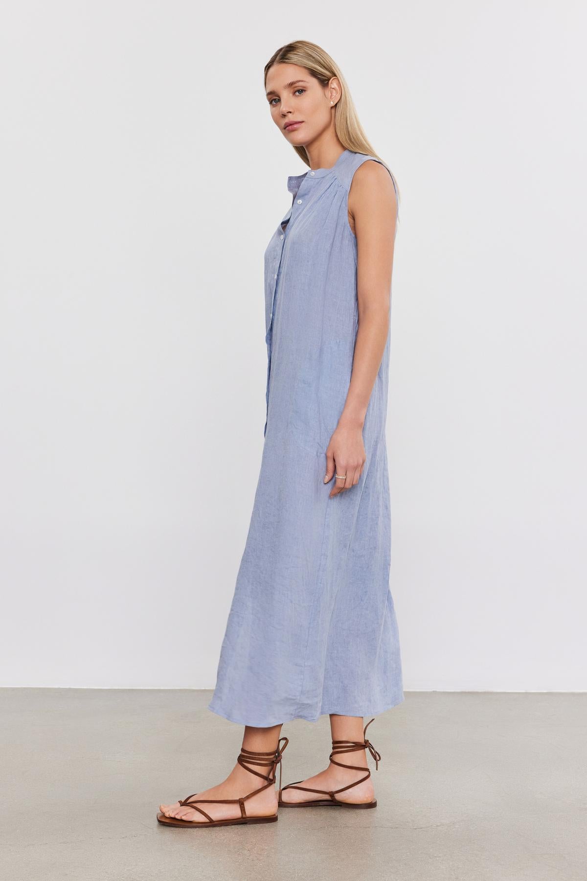   A woman wearing a sleeveless, light blue Velvet by Graham & Spencer Luna Linen Dress and brown strappy sandals stands facing sideways on a plain background. 