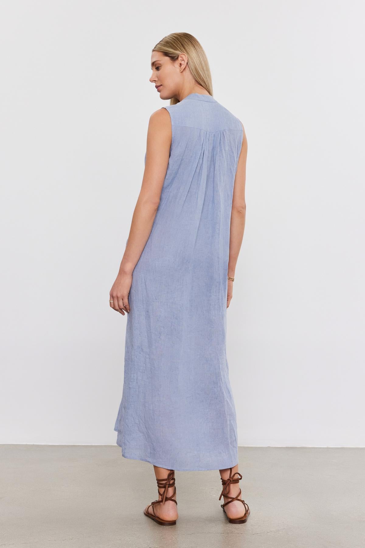   A woman in a Velvet by Graham & Spencer LUNA LINEN DRESS sleeveless maxi-length dress and brown lace-up sandals, standing against a white background, viewed from the side. 