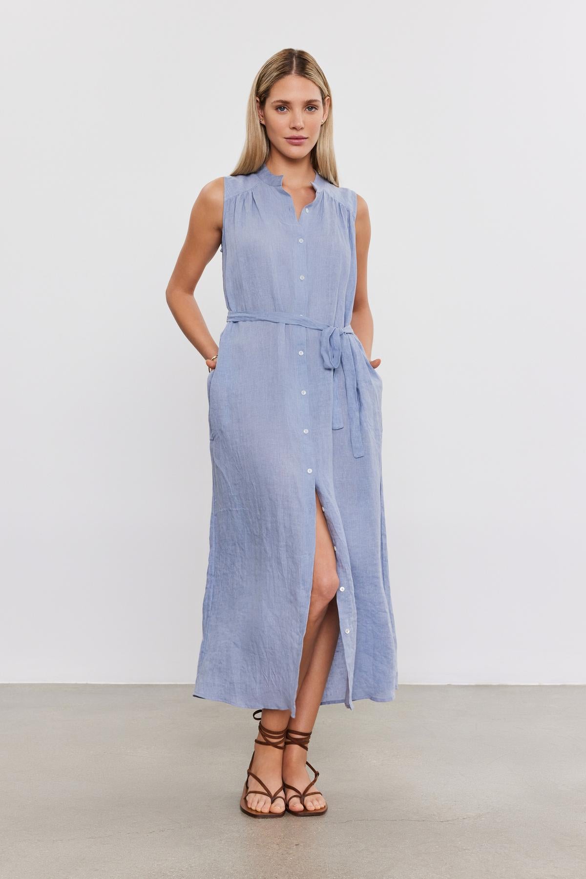   Woman standing in a studio, wearing a light blue sleeveless LUNA LINEN DRESS by Velvet by Graham & Spencer with a button front, waist tie, and high slit, paired with brown sandals. 