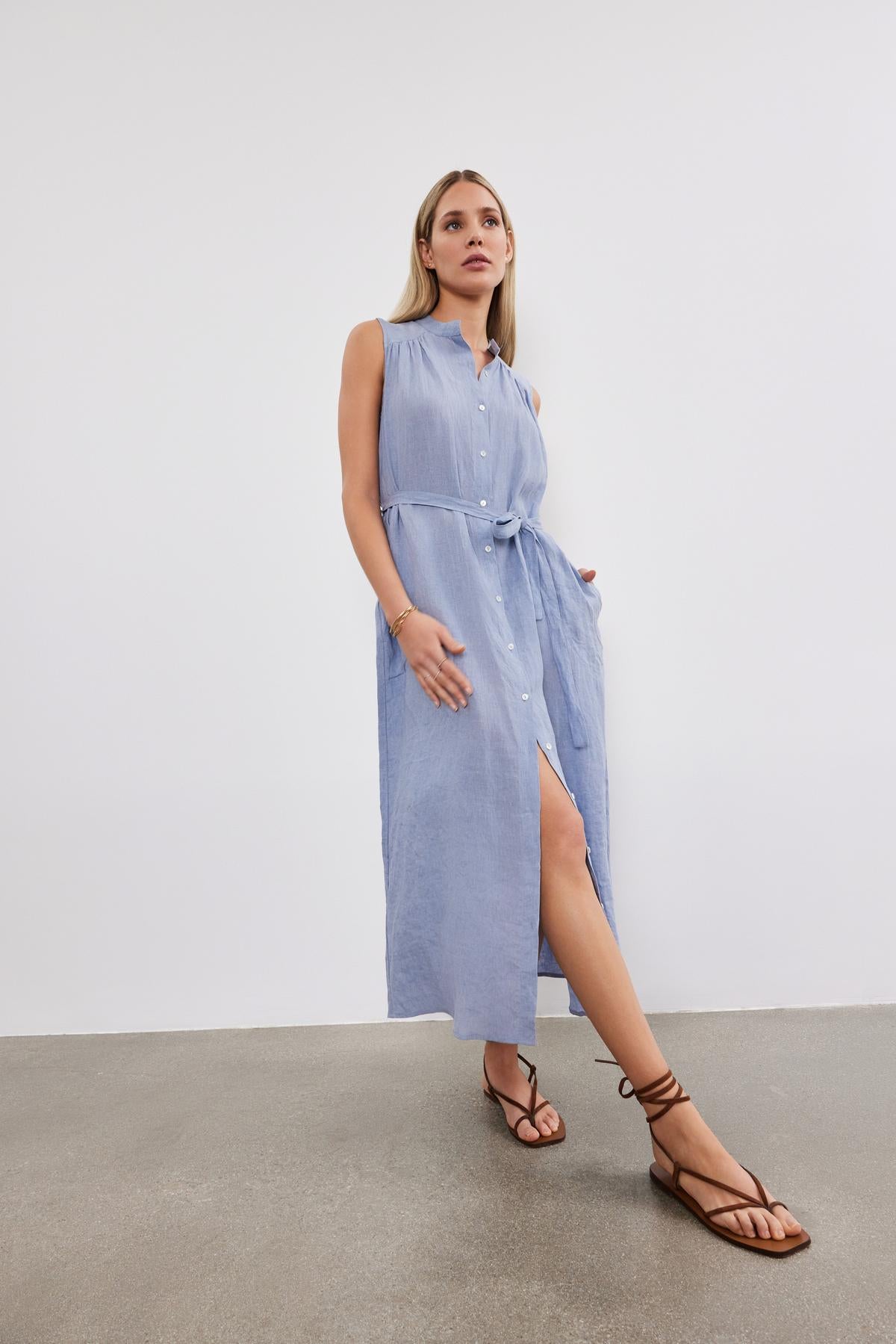   A woman stands in a studio, wearing a Velvet by Graham & Spencer LUNA LINEN DRESS with a slit, accessorized with brown sandals. 