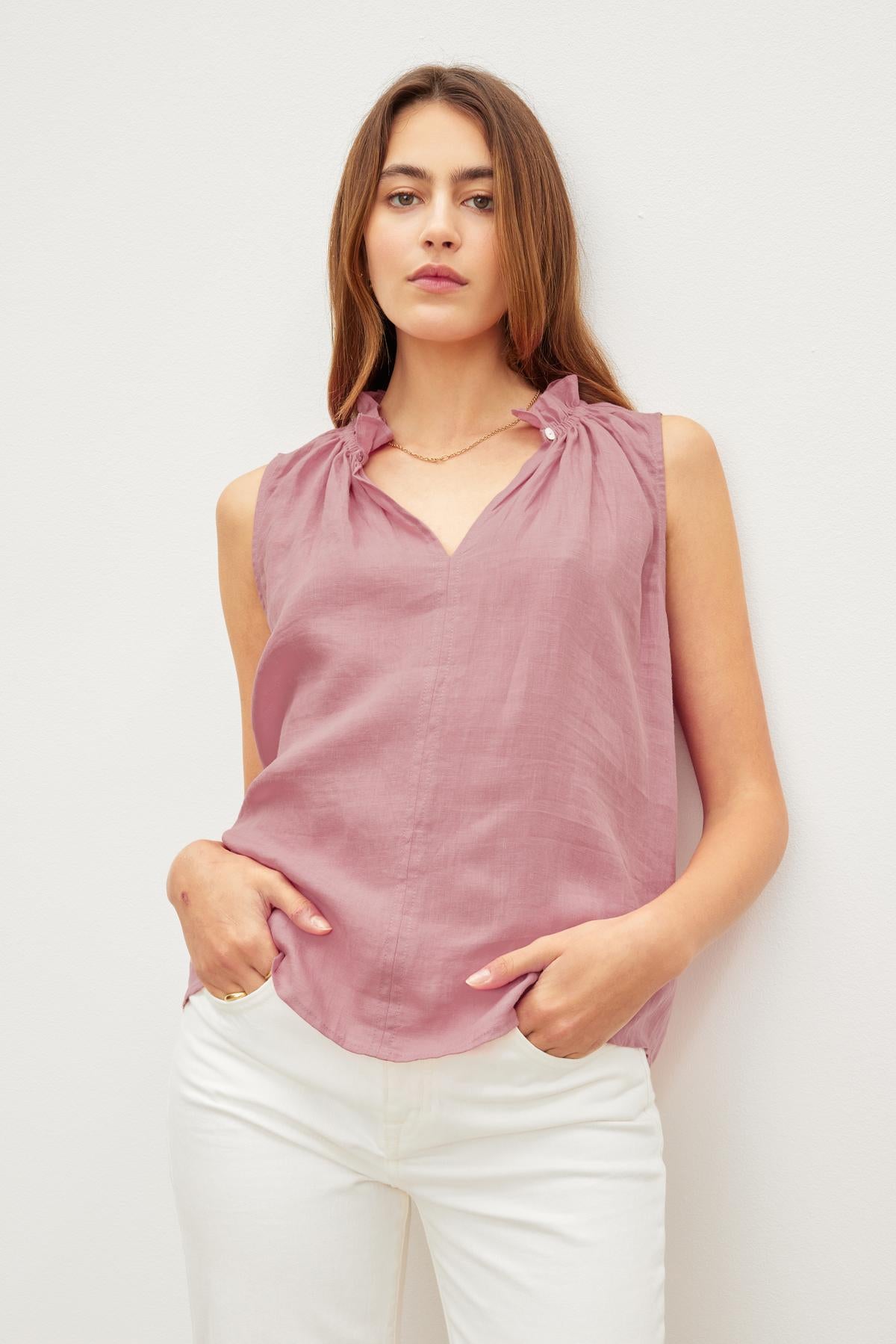   Woman in a pink Velvet by Graham & Spencer MASIE LINEN TANK TOP and white pants standing against a white background, hands in pockets, looking at the camera. 