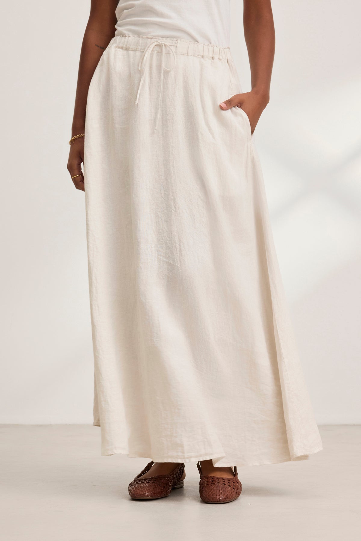 A person stands against a light background, wearing the long MEMPHIS LINEN SKIRT by Velvet by Graham & Spencer, featuring a cream color, woven linen fabric, elastic waist, and pockets. It is paired with brown shoes.-38819723280577