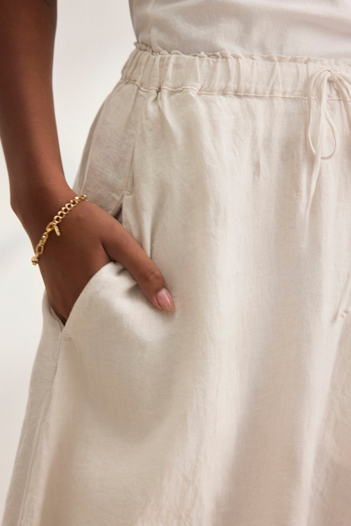 Close-up of a person wearing the MEMPHIS LINEN SKIRT by Velvet by Graham & Spencer, featuring an elastic waist and matching top. Their hand rests in the skirt pocket, highlighting a gold bracelet.-38819723706561