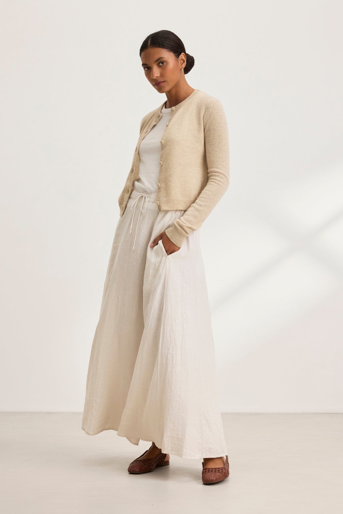 A person stands against a plain backdrop wearing a beige cardigan, white top, and the Velvet by Graham & Spencer MEMPHIS LINEN SKIRT.-38819723018433