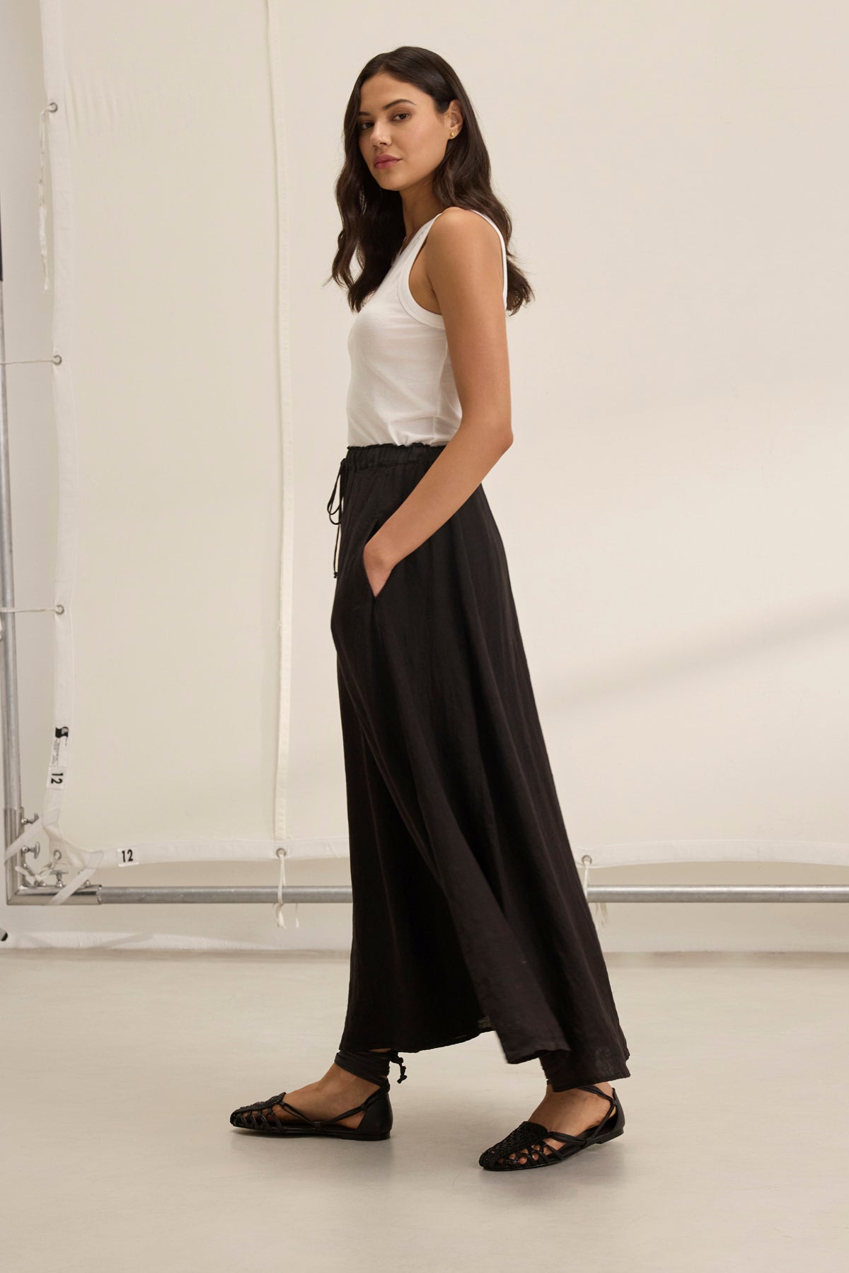 A woman stands sideways, her silhouette elegant in a white sleeveless top and the Memphis Linen Skirt by Velvet by Graham & Spencer, paired with sleek black flats.-38819723182273