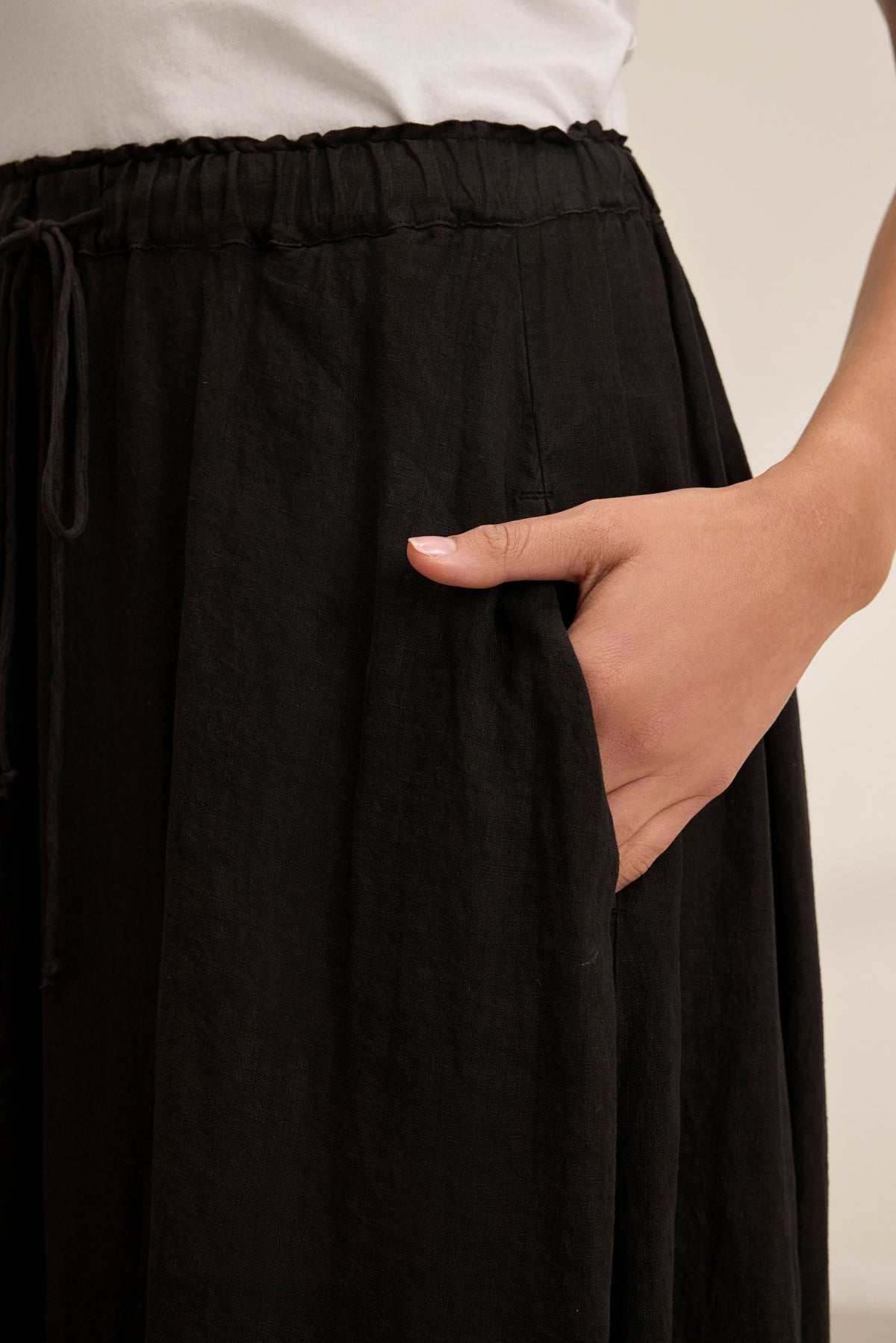 Someone casually slips their right hand into the pocket of a black MEMPHIS LINEN SKIRT with an elastic waist by Velvet by Graham & Spencer, paired with a white top.-38819723804865