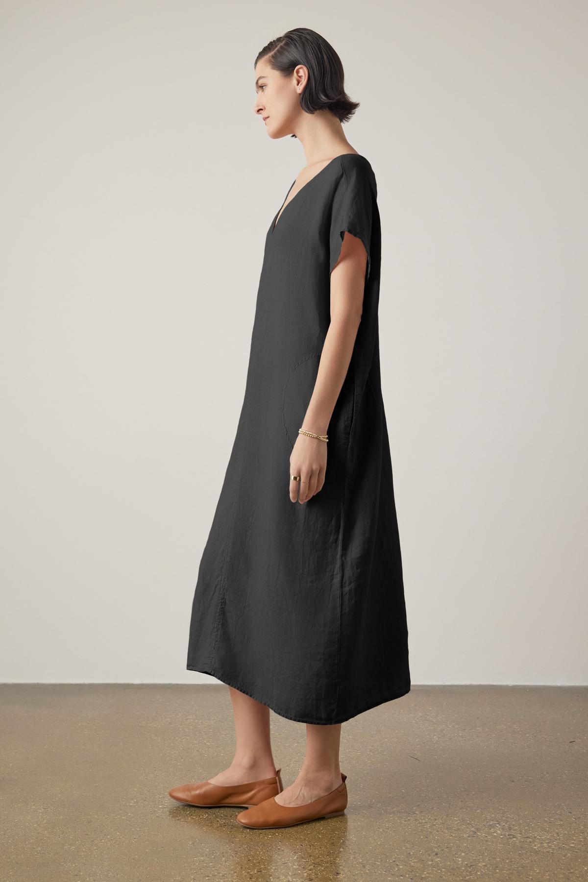 A person with short hair is standing sideways, wearing a loose-fitting, ankle-length black MONTANA LINEN DRESS by Velvet by Jenny Graham and brown flat shoes, against a plain white background.-36863315443905