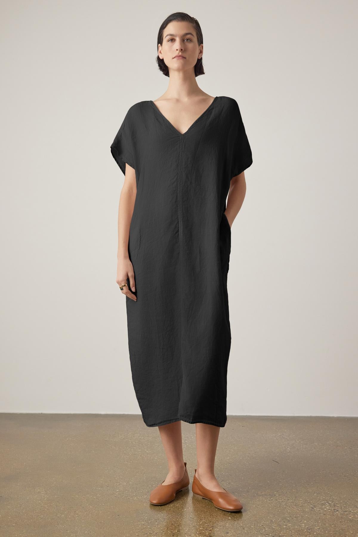   A woman in a loose black Montana linen dress by Velvet by Jenny Graham with a V neckline and brown loafers stands against a neutral backdrop, looking directly at the camera. 