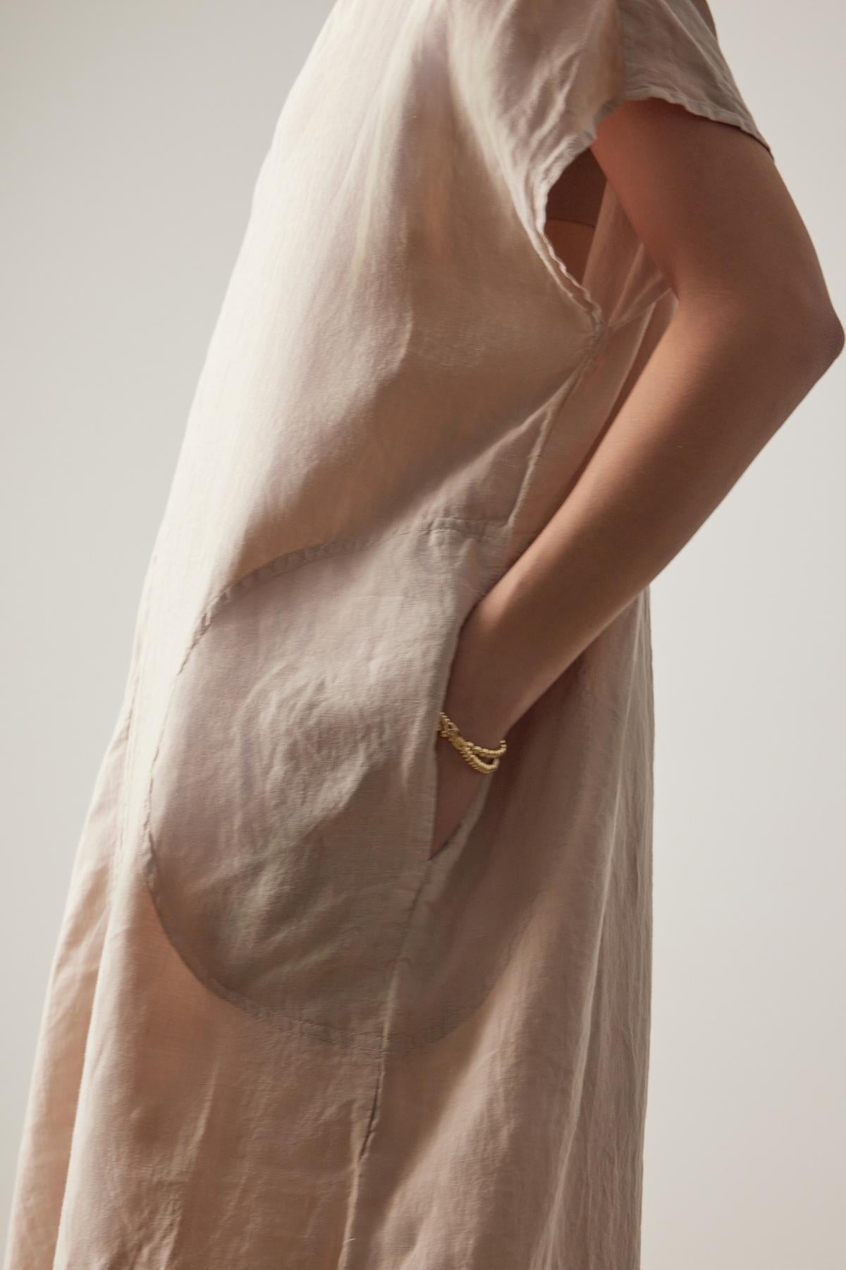   A close-up of a person in a Velvet by Jenny Graham Montana linen dress with a large pocket, highlighting a detail of a gold bracelet on their arm. 