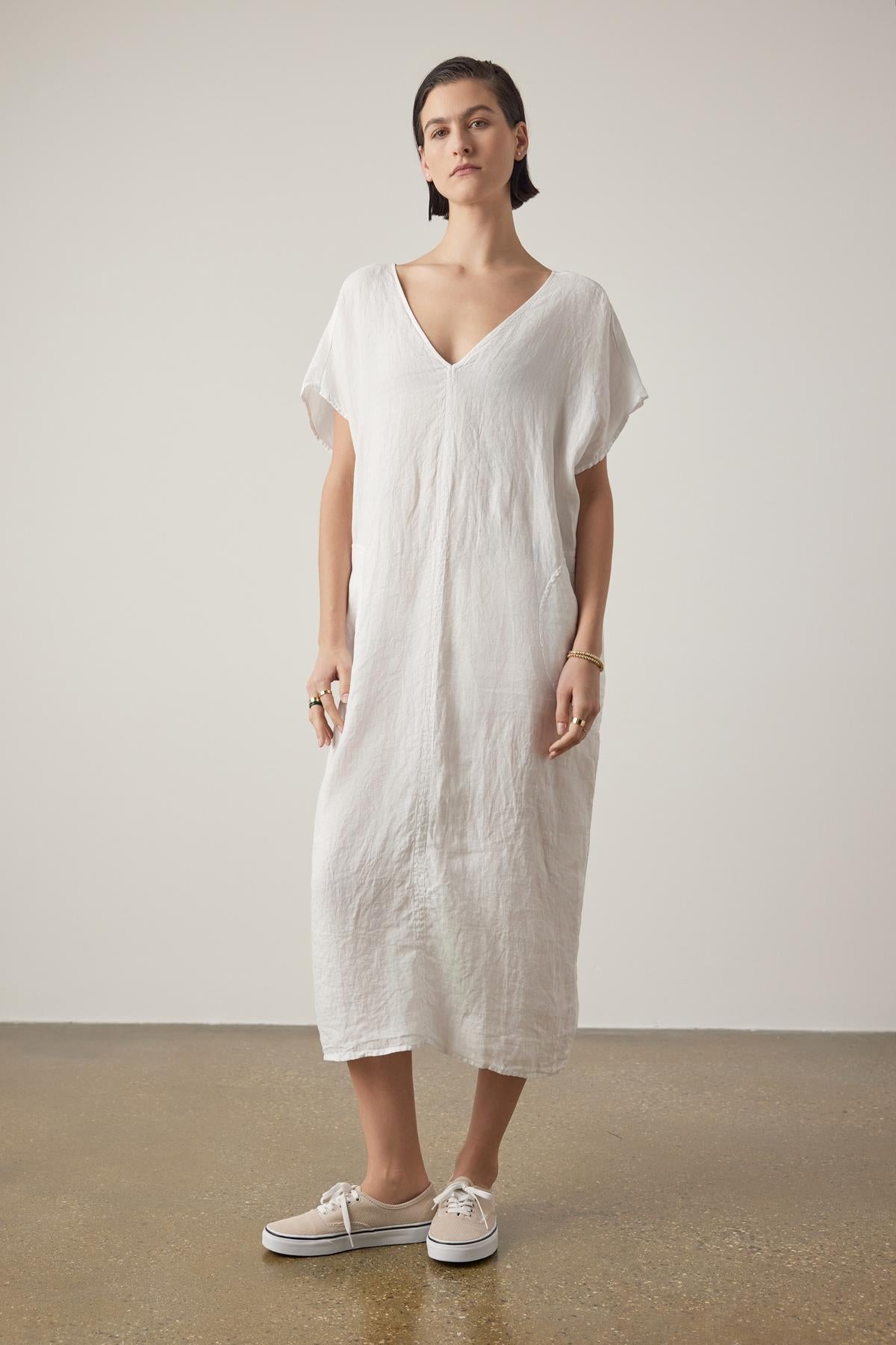   Person standing in a minimalist setting, wearing the MONTANA LINEN DRESS by Velvet by Jenny Graham and white sneakers. They have short hair and are looking directly at the camera with a neutral expression. 